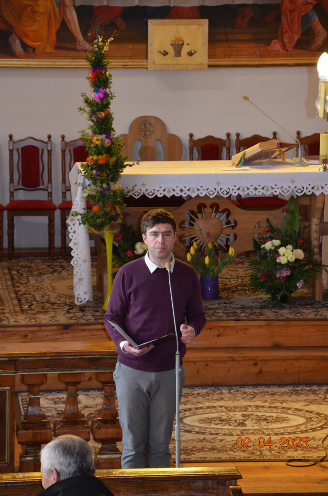 Główny prowadzący przegląd chóru Pan Mariusz czerwiński przedstawia chóry przez mikrofon. w tle ołtarz i palma wielkanocna. 