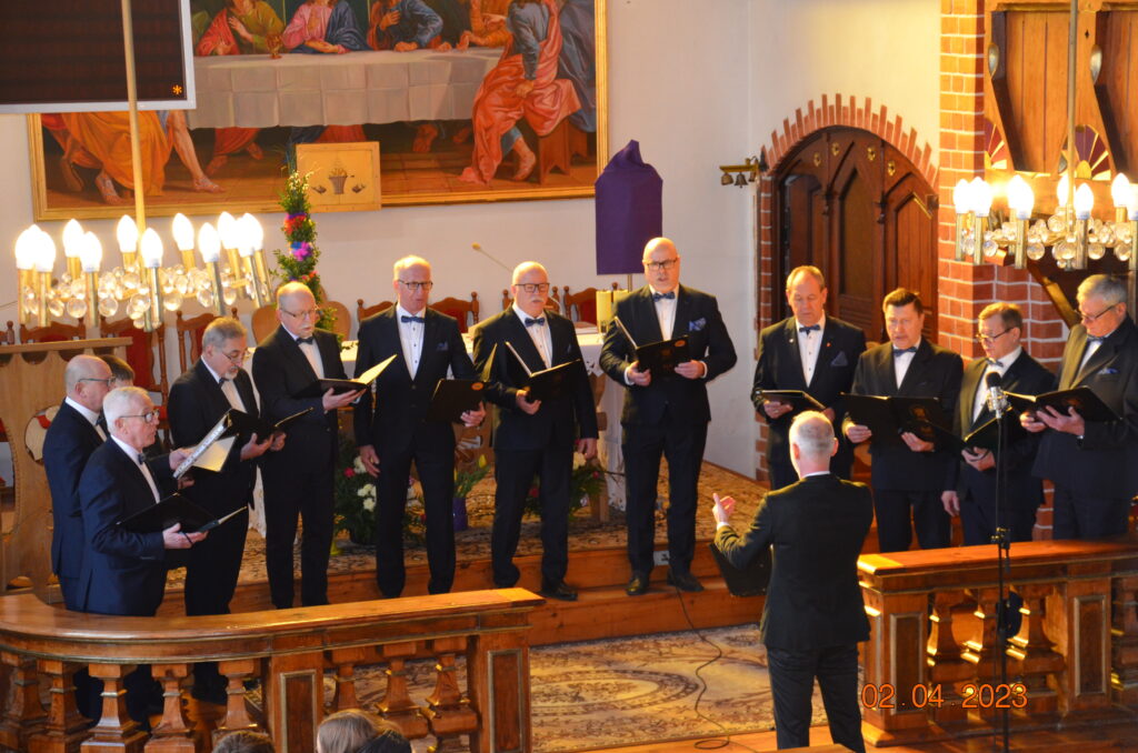 grupa 13 mężczyzn ubranych w czarne garnitury i białe koszule śpiewają pieśni wielkopostne. 