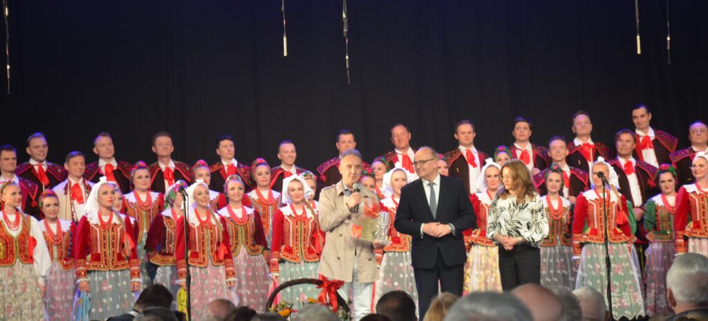 koncert pieśni i tańca Śląsk.  w tle tancerze ubrani w kolorowe stroje. przed mikrofonem dyrektor zespołu oraz wójt gminy trąbki wielkie 