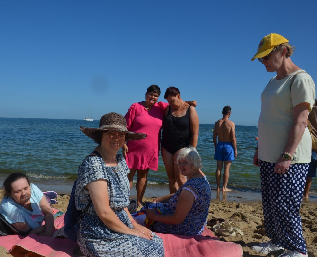 morze Bałtyckie. Błękitne niebo, niebieska woda, na plaży na kocu siedzą trzy Panie , po środku Pani Justyna w brązowym kapeluszu, nad Nimi pozują do zdjęcia Pani Basia w czarnym stroju kąpielowym oraz Pani paulina w różowej sukience, 