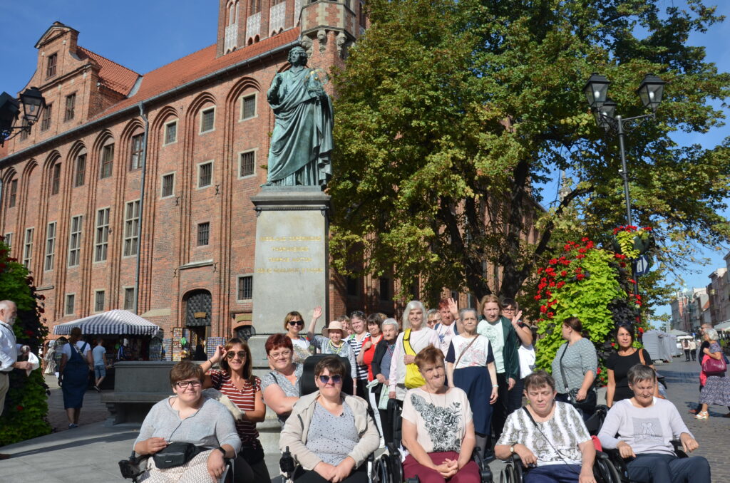 Miasto Toruń Słoneczny dzień, Pomnik Mikołaja Kopernika, spora grupa pań pozuje do zdjęcia, uśmiecha się. 