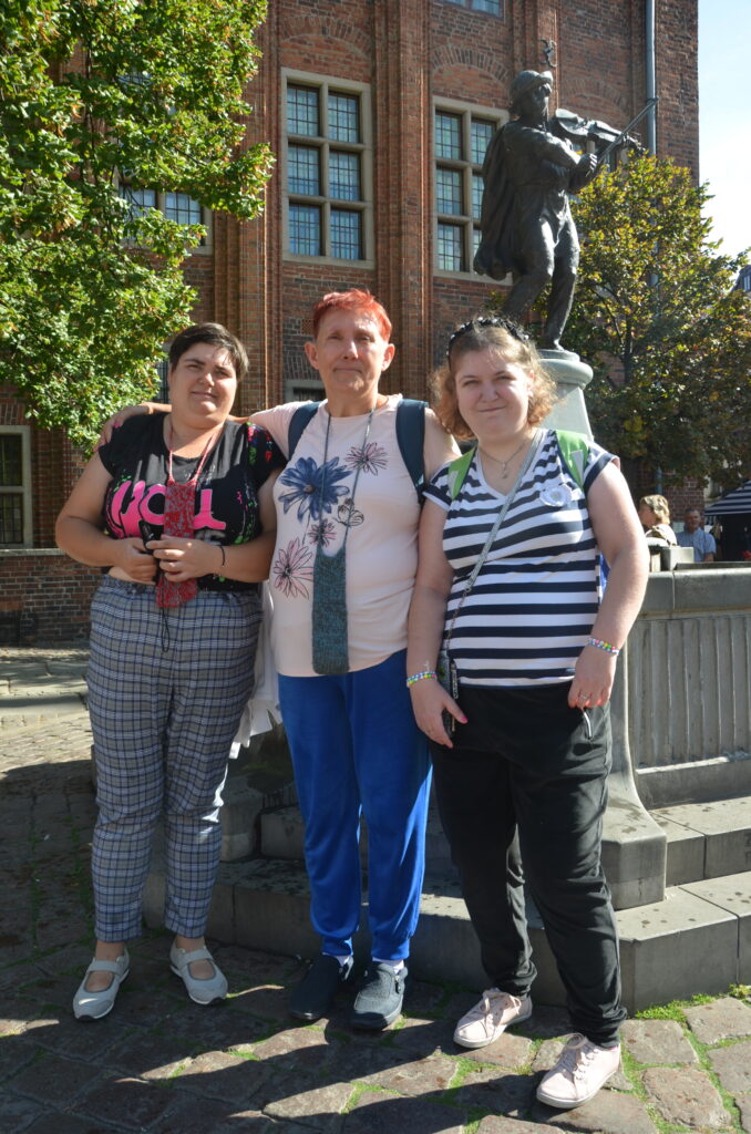 Toruń. słoneczny dzień. Trzy panie pozują do zdjęcia przy pomniku chłopca ze skrzypcami 