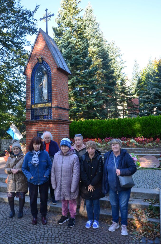 pielgrzymka do Gietrzwałdu.
Grupa siedmiu pań stoi przed kapliczką objawienia Marki Bożej. Uśmiechają się do zdjęcia. w tle błękit nieba, zielone drzewa