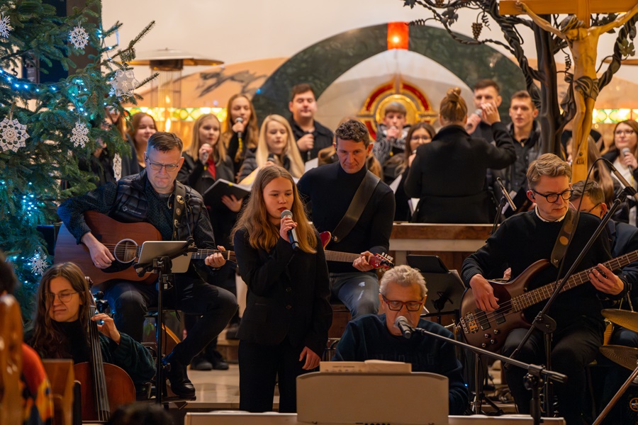 koncert kolęd. w tle spora grupa muzyków grających na instrumentach, na środku stoi młoda dziewczynka z mikrofonem, wykonuje śpiew 