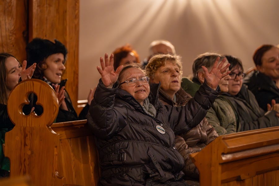 Koncert kolęd. Grupa pięciu pań siedzi w ławkach kościoła, wykonuje radosne uniesienie rąk w górze, uśmiecha się 