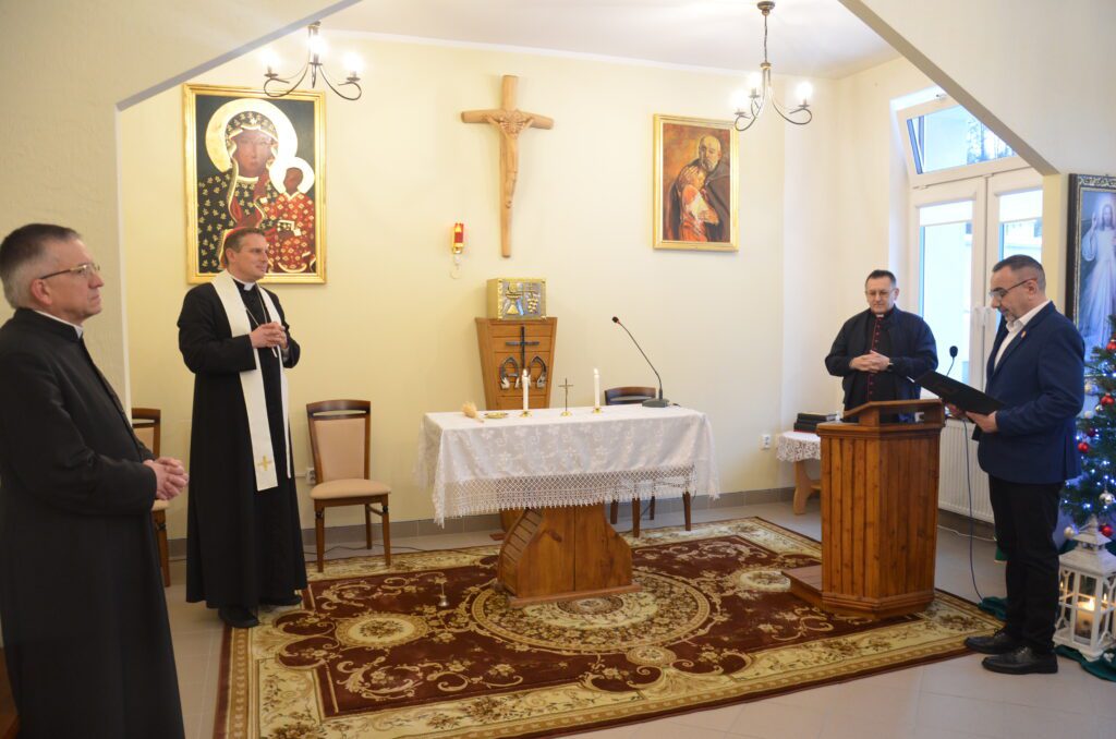 Kaplica W Domu. Główny ołtarz , na środku ołtarza biały obrus, dwie zapalone świece  , krzyż . przy mównicy mówi do mikrofonu Pan Dyrektor , wiata biskupa . po obu stronach ołtarza stoi dwóch księży. 