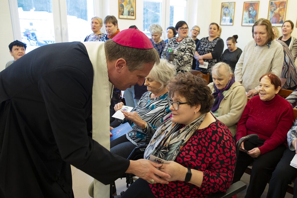 w Kaplicy. Biskup pochyla się i rozmawia z mieszkanką siedzącą na wózku, wręcza jej obrazek święty. 