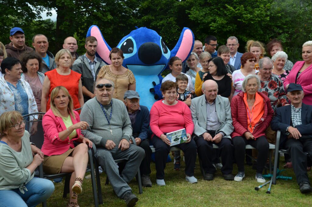zdjęcie grupowe na tle zielonych drzew Stitch postać bajkowa w centralnym miejscu, wokół uczestnicy ,goście pracownicy część siedzi na krzesłach