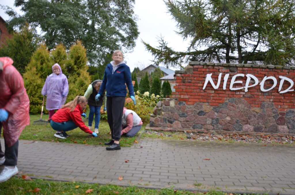 inny pomnikwidac grupke ludz jedna kobieta soi na chodniku i się patrzy na fotografa, w oddali widać druga panią stojącą i trzymajacą grabi 2 osoby są schylone sadzą kwiaty