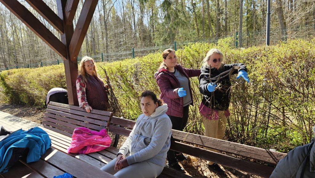 pod wiatami, ławki na jednej siedzi kobieta , za nią widać 3 panie robiące coś przy żywopłocie