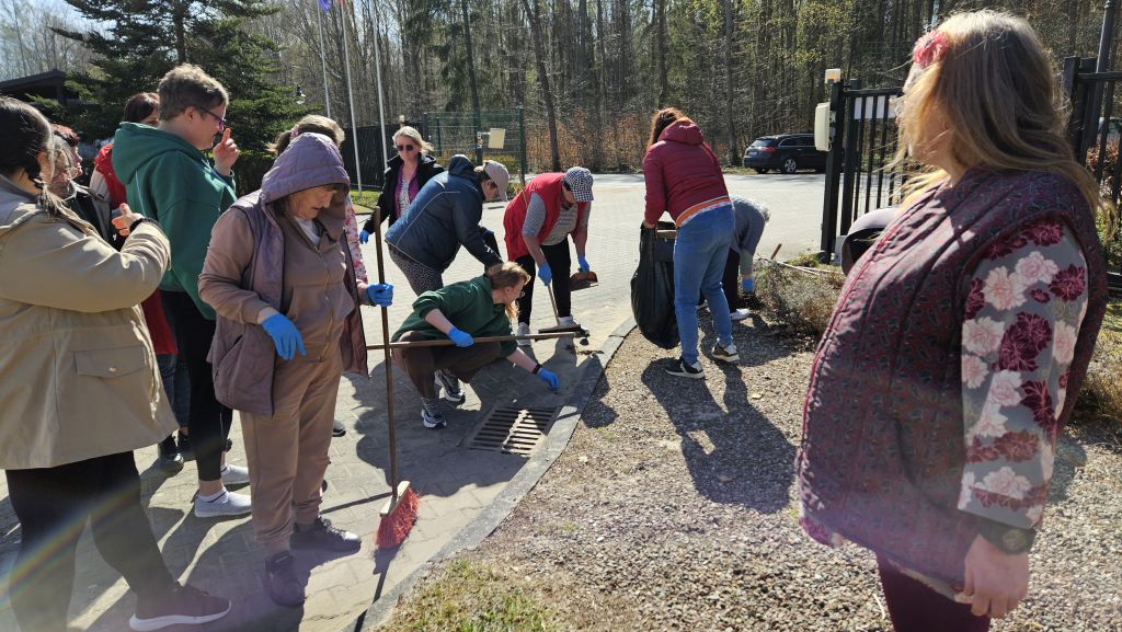 na powietrzu przed bramą otwartą krzątają się znaczna grupa kobiet, widać miotłę, grabie , chaczke , część jest pochylona, część stoi