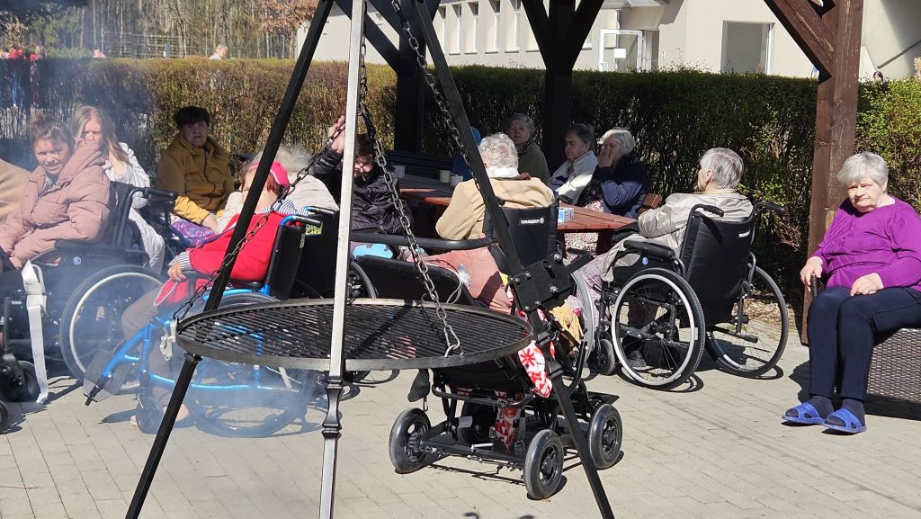 pod wiatami widać siedzace panie część na wózkach i reszta na ławkach, na pierwszym planie jest widoczne palenisko i lecący dum z niego