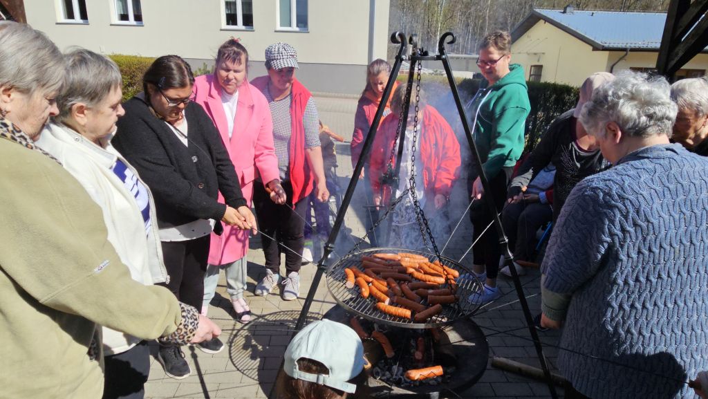 pod wiatami widać palenisko na rusztcie widać duzo kiełbasek, wokół niego stoi dużo pań ktore pieką sobie kiełbaski