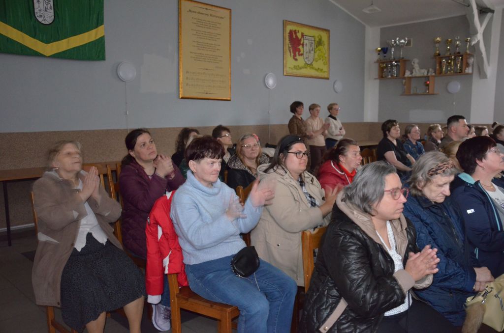 następne zdjęcie pubiczności siedzą panie i biją brawa, na ścianach wiszą obrazy obok półka z pucharami 