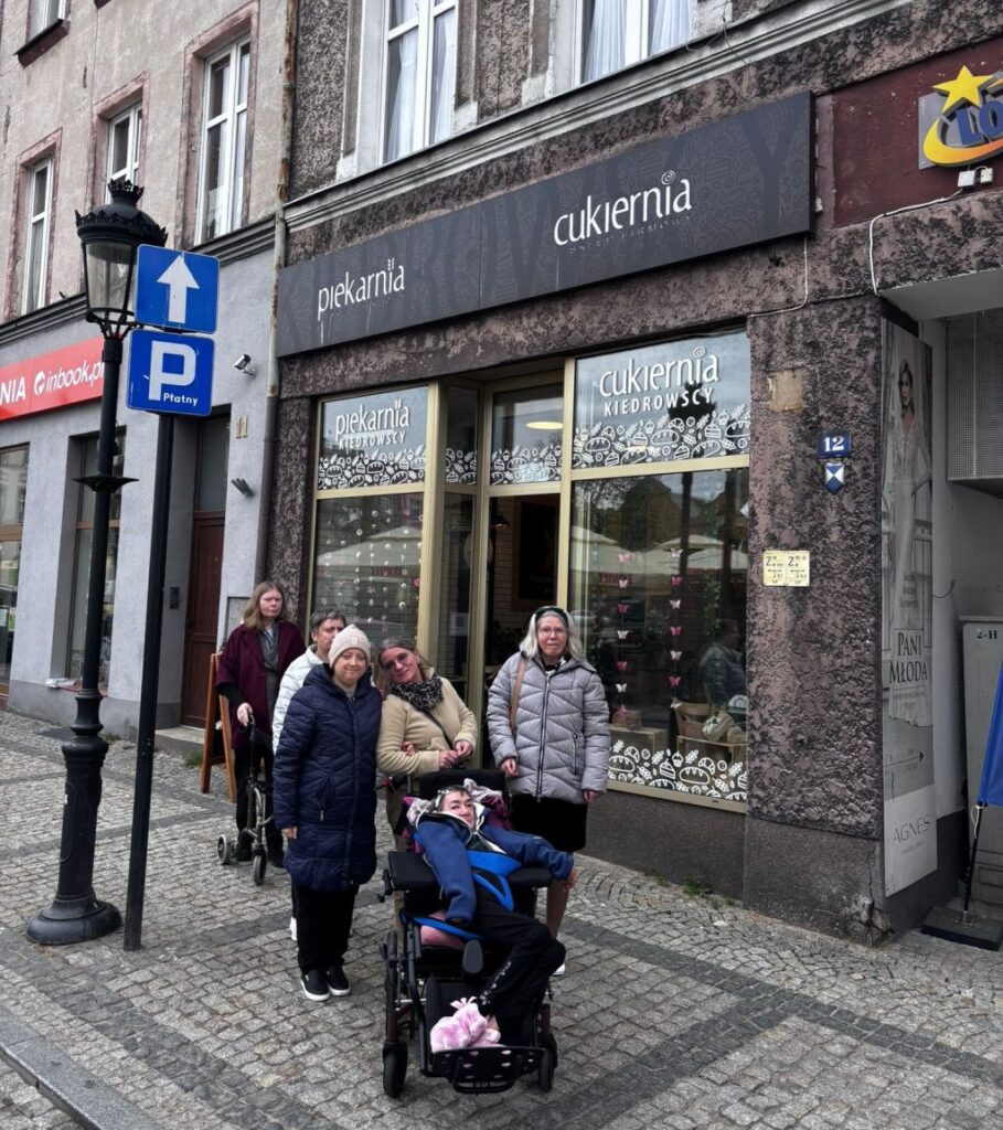 grupa tych samych pań stoi przed cwitryną cukierni i pozują do zdjęcia, widać  boku piękną lampę stojącą