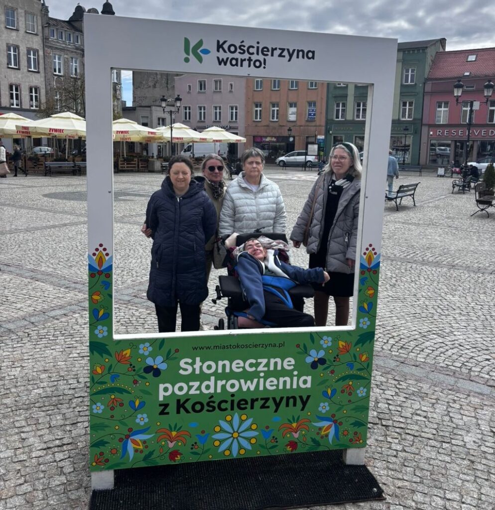 panie stoją na rynku  w kościerzynie, jest wyłożony kamieniem brukowym w kolorze szarym, stoi 4 kobiety i jedna jest na wózku, ramka w której stoją jest ozdobiona wzorem kaszubskim i u góry pisze ,, warty kościerzryna'' , w dali widać parasole przeciwsłoneczne i stliki, wokółplacy stoją kamienice 