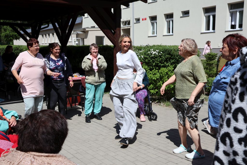 wiata ogrodzona rzywopłotem, widać budynek w tle w centrum zdjęcia są 6 kobiet tańczących i uśmiechnięte