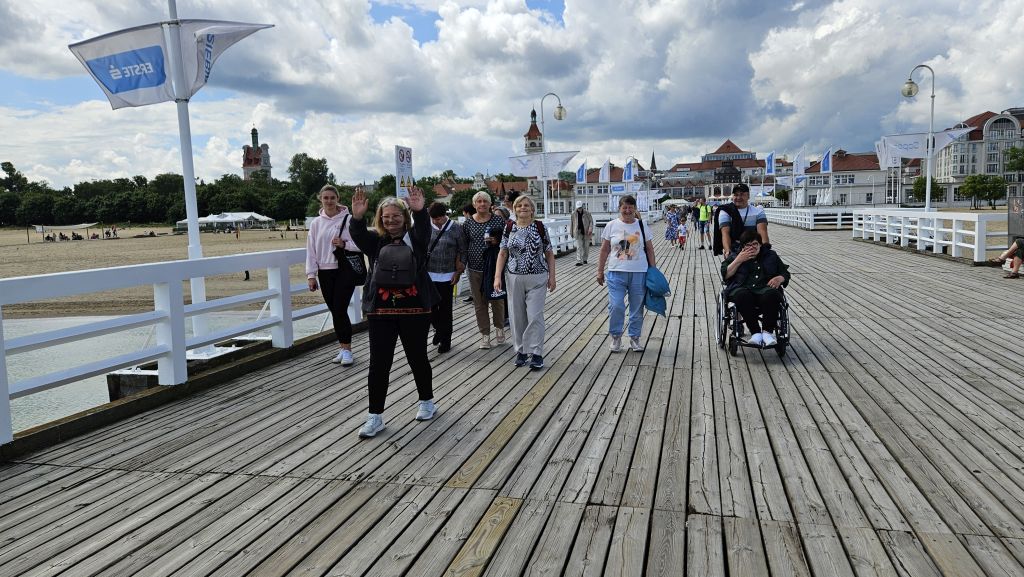 sopot molo , panie idą grupą po molo widać deski , balustrady, jedna macha uśmiechnięta, inna jedzie na wózku