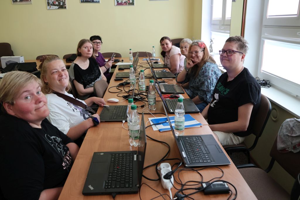 pomieszczenie 8 kobiet siedzi przed laptopami i uśmiecha się do fotografa, na stole widać również butelki z wodą