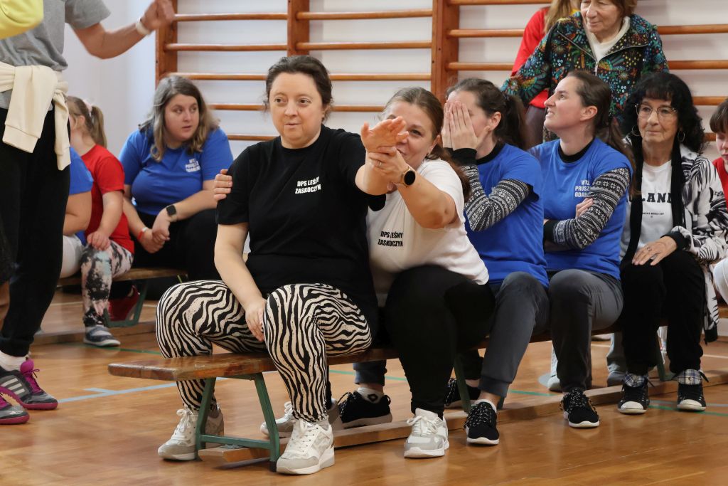 inne miejsce inne panie jedna w czarnej koszulce a druga w białej siedz jedna za druga a za nimi inne uczestniczki 2 panie w niebieskich koszulkach, widac jeszcze kilka osob w tle