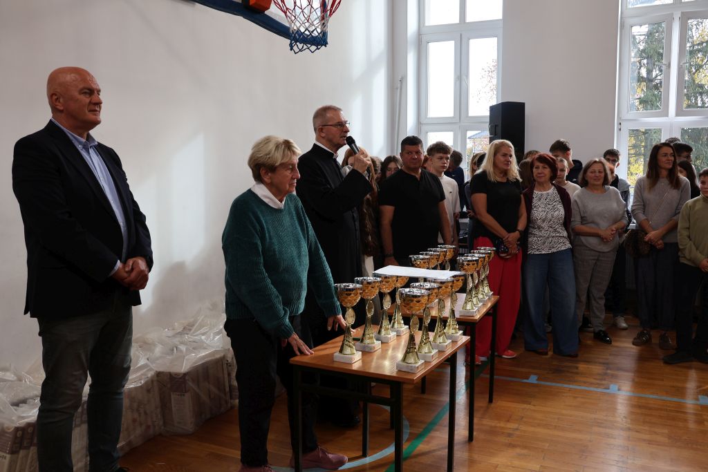 grupa osób 3 stoja przed stołem z pucharami ksiądz trzyma mikrofon i przemawia , panie w zielonym swetrze opiera się o stół